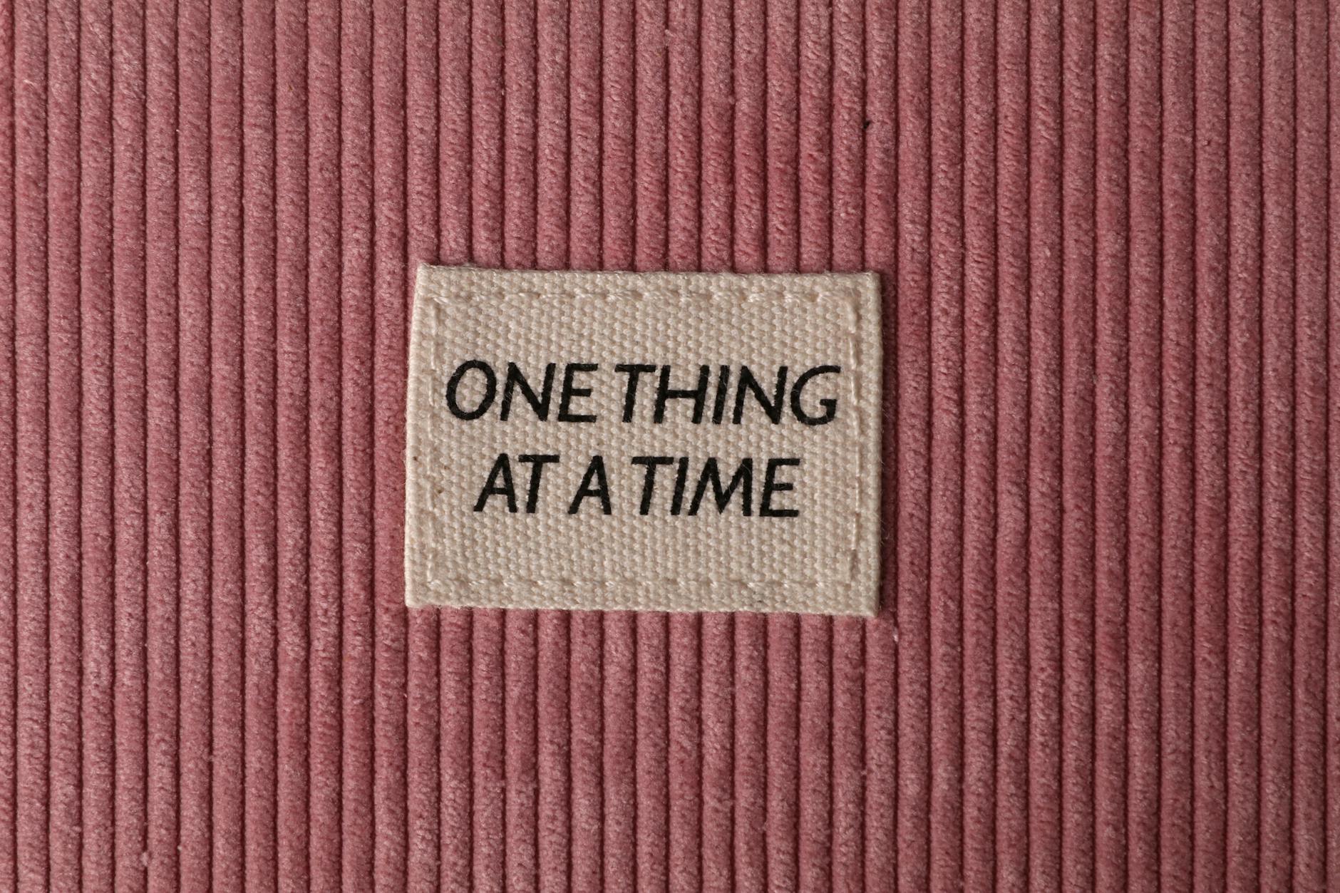 Metka z napisem One thing at a time na różowym materiale sztruksowym